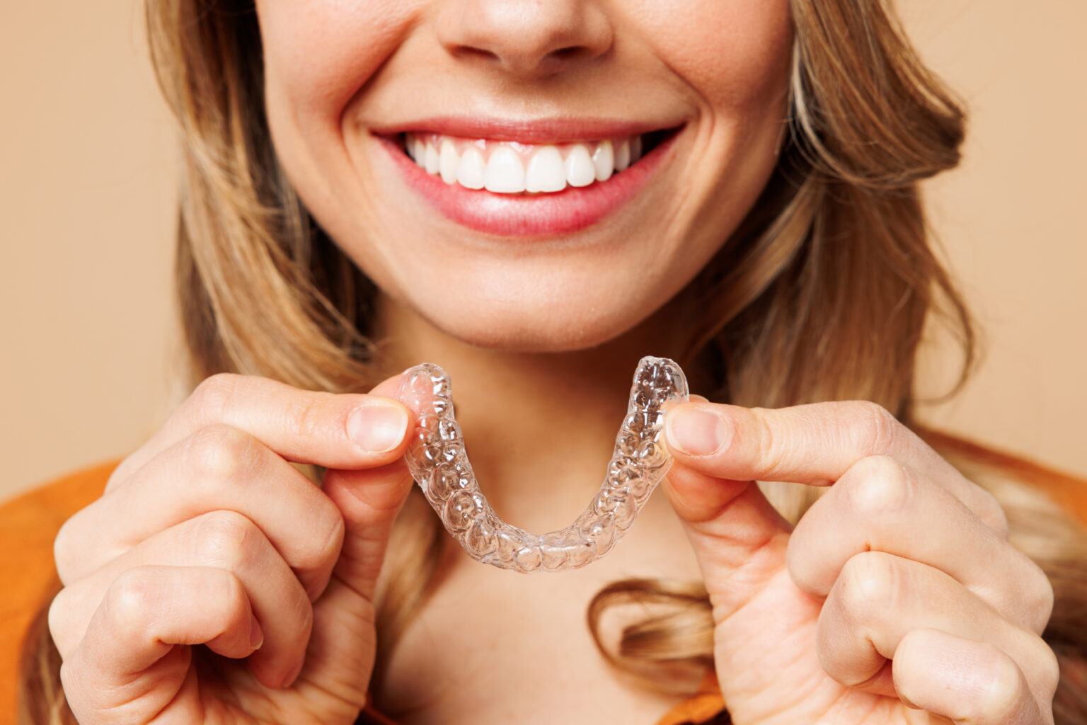 Close up cropped young woman wear orange shirt casual clothes hold in hand invisible transparent aligners, invisalign bracer smile isolated on plain pastel light beige background. Lifestyle concept